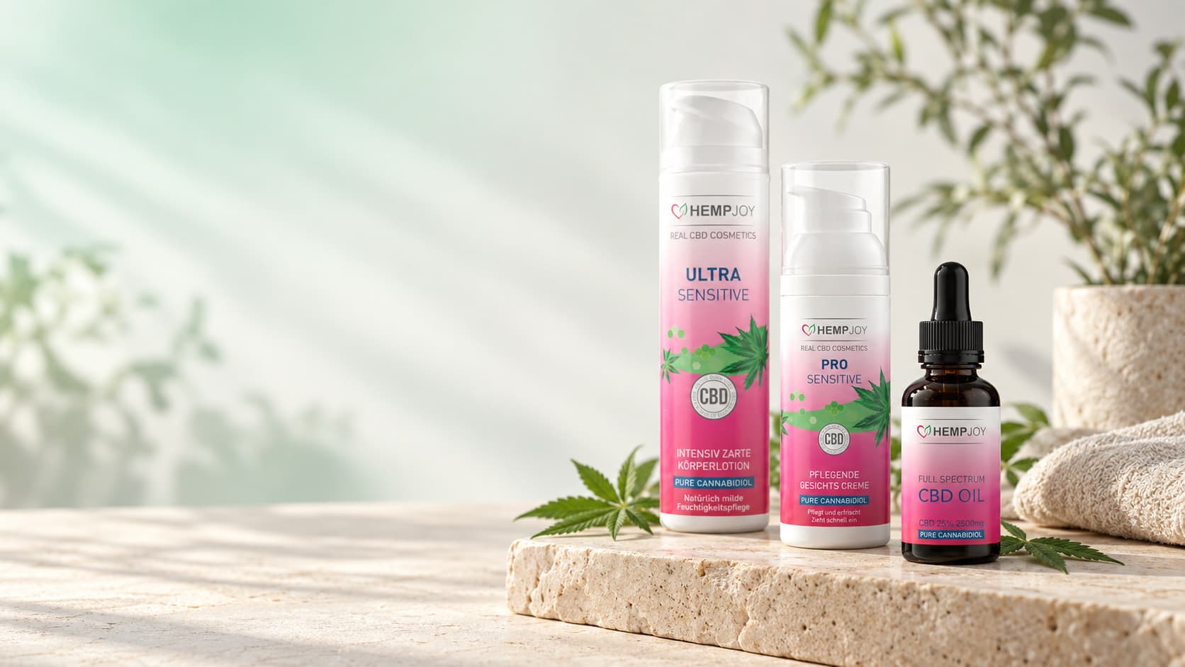 Drei HempJoy-Produkte auf einer hellen Steinplatte: Körperlotion, Gesichtscreme und eine Tropfflasche CBD-Öl, daneben Hanfblätter und ein gefaltetes Tuch